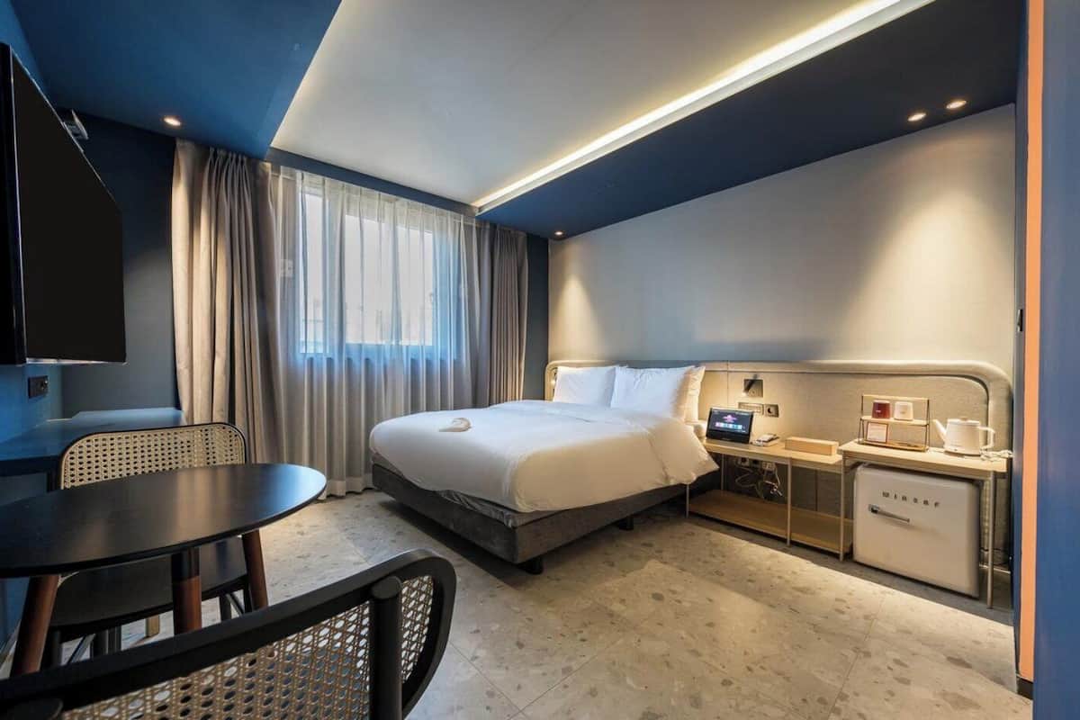 Habitación de hotel con cama, escritorio, silla, televisor, mesita de noche con lámpara y minibar.