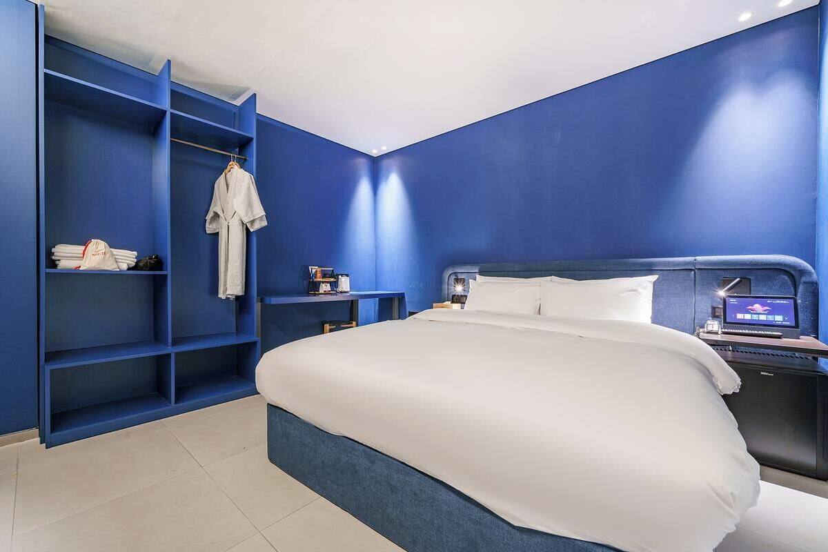 Una habitación de hotel moderna con una cama grande, un armario con estantes, un escritorio con una computadora y una mesita de noche.