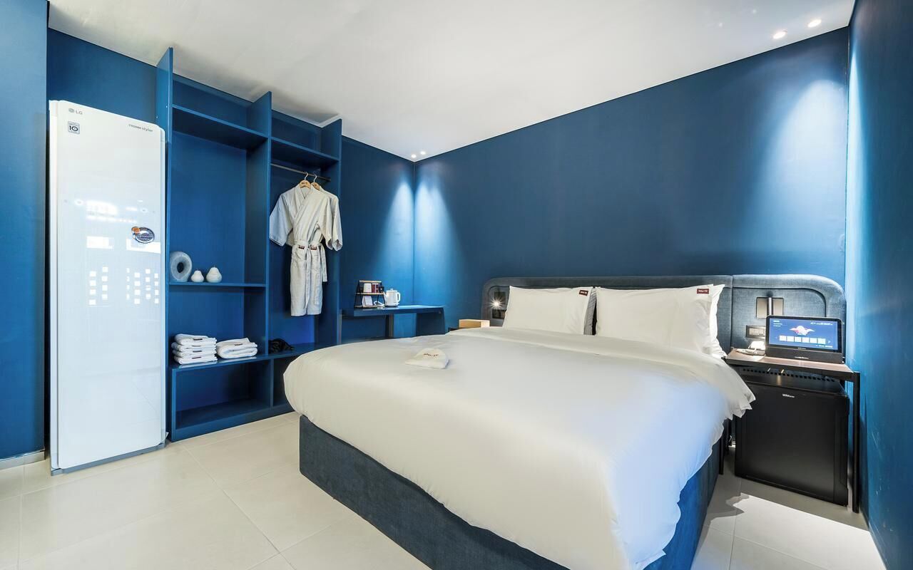 Una habitación de hotel moderna con una cama grande, televisión integrada, mini refrigerador y una unidad mural azul con estantes.