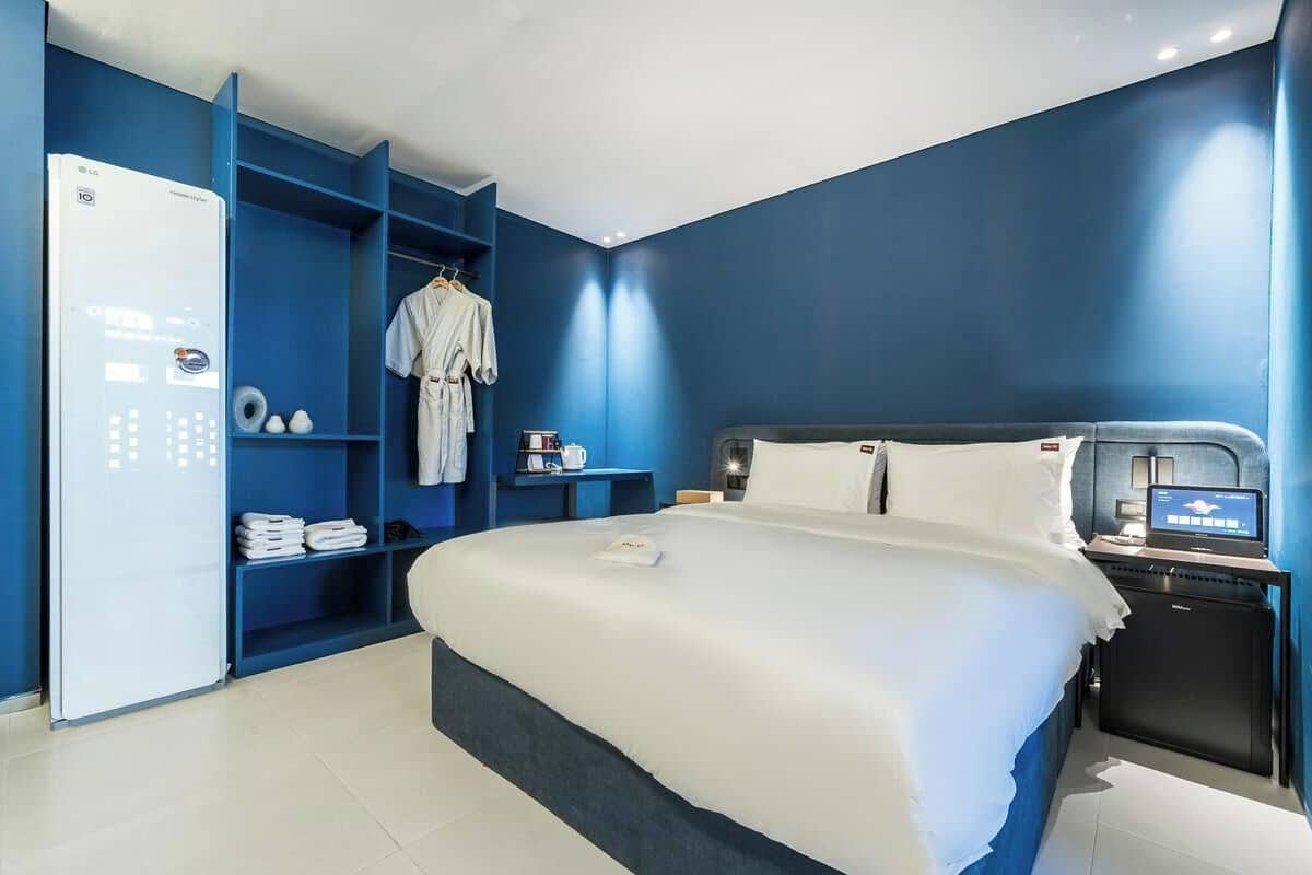 A modern hotel room with a large bed, a built-in TV, a mini-fridge, and a blue wall unit with shelves.
