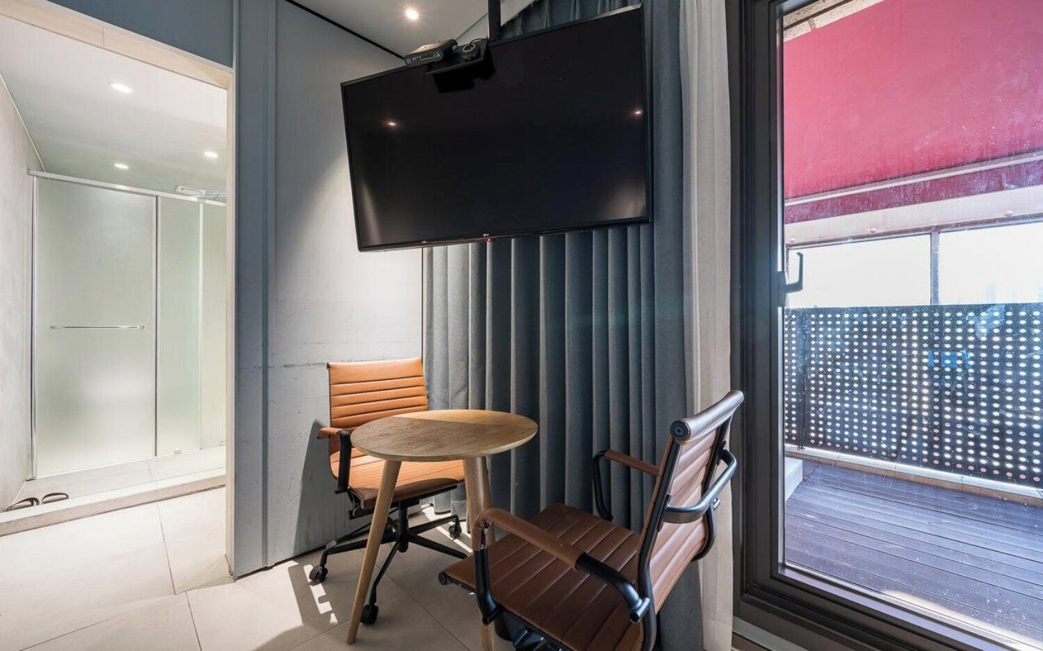 A modern room with a flat-screen TV, two chairs, a small round table, and a sliding glass door leading to an outdoor balcony.