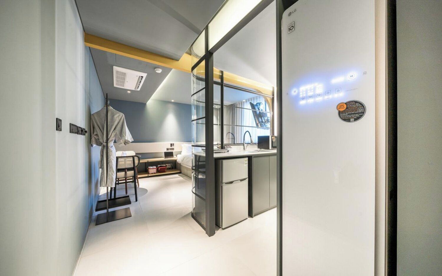 Una habitación de hotel moderna con kitchenette, un espejo grande y una pared divisoria de vidrio.