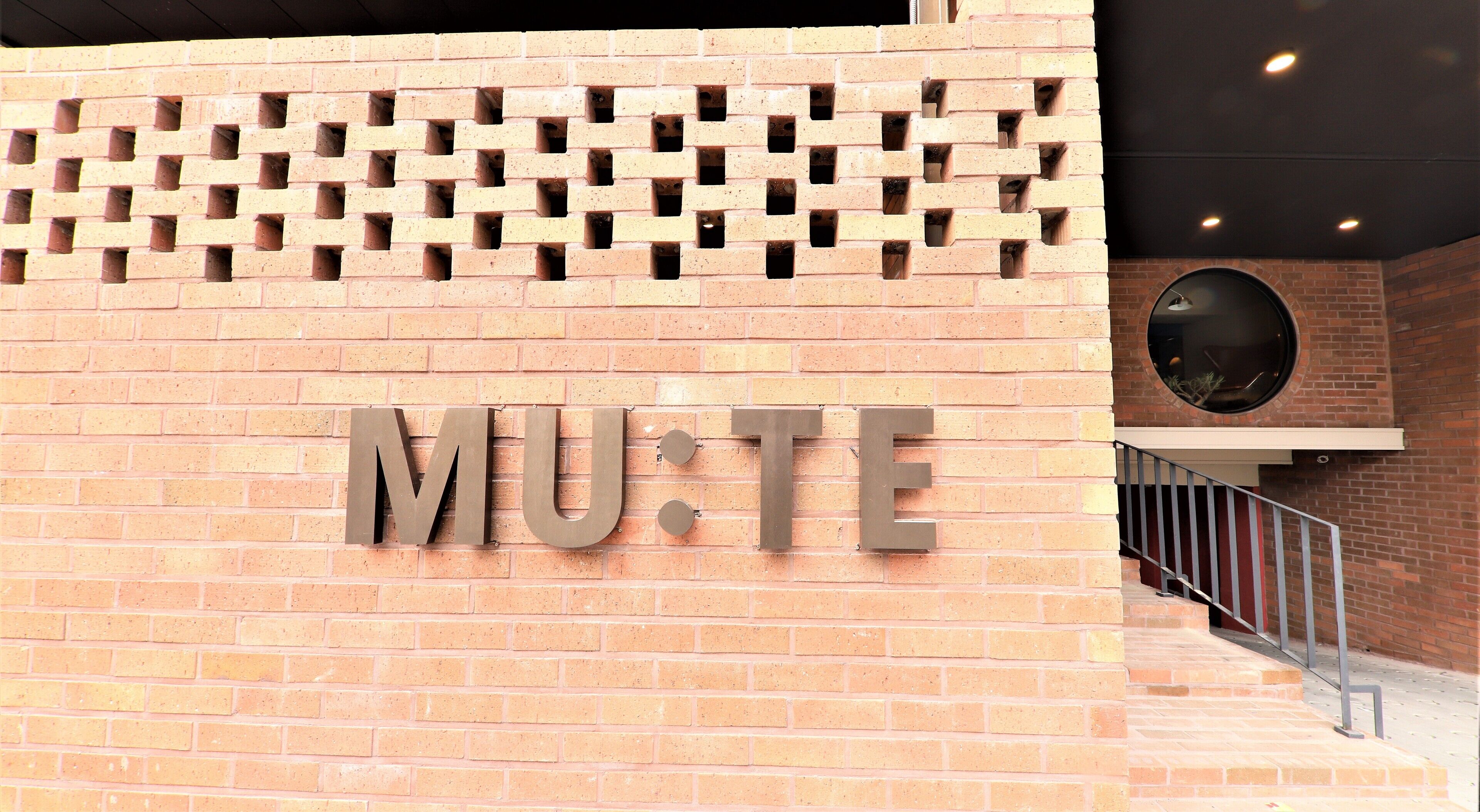 Un muro de ladrillo con la palabra "MU:TE" en letras grandes, una ventana circular y una barandilla de metal.