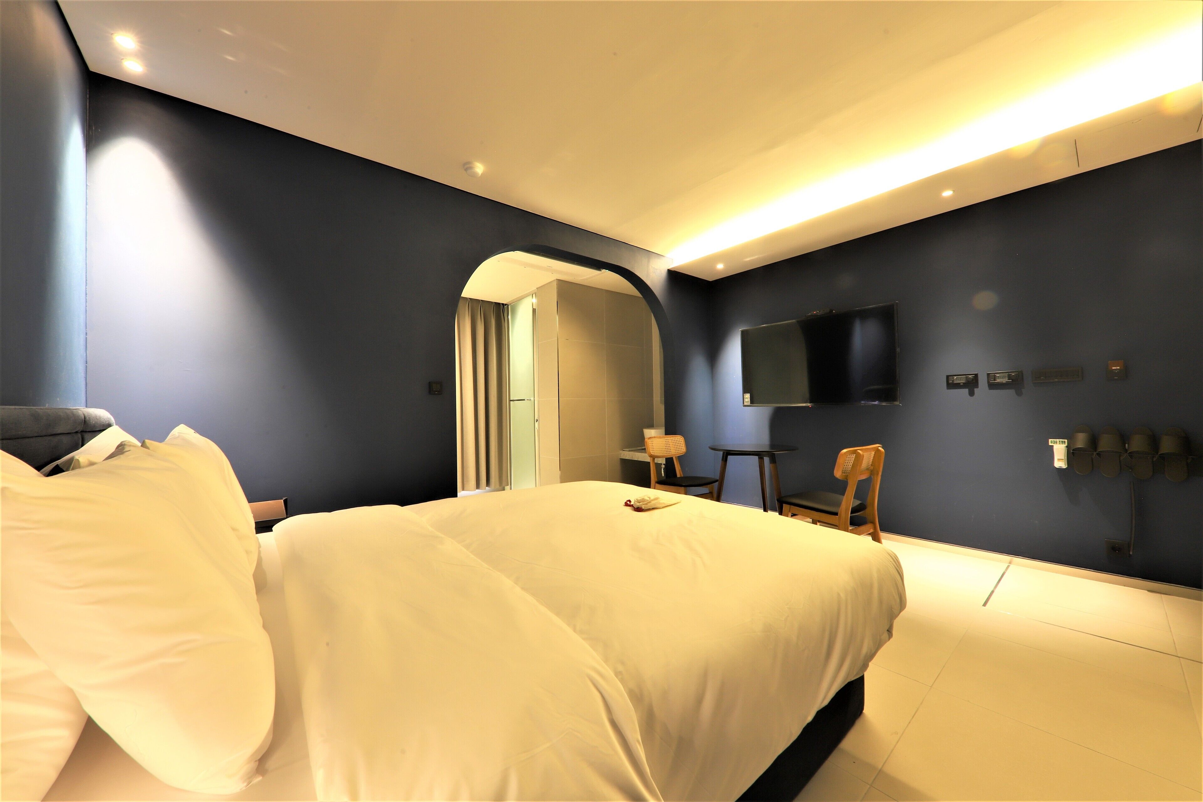 A modern hotel room with a large bed, a desk, a chair, and a flat-screen TV mounted on the wall.