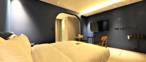 A modern hotel room with a large bed, a desk, a chair, and a flat-screen TV mounted on the wall.