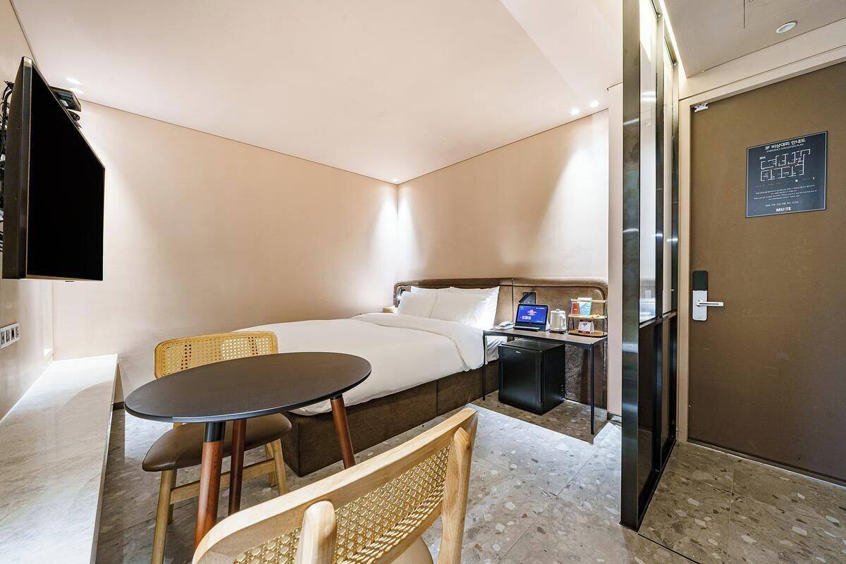 Habitación de hotel con cama, una mesita, una silla, un escritorio con una computadora portátil y una puerta con un cartel.