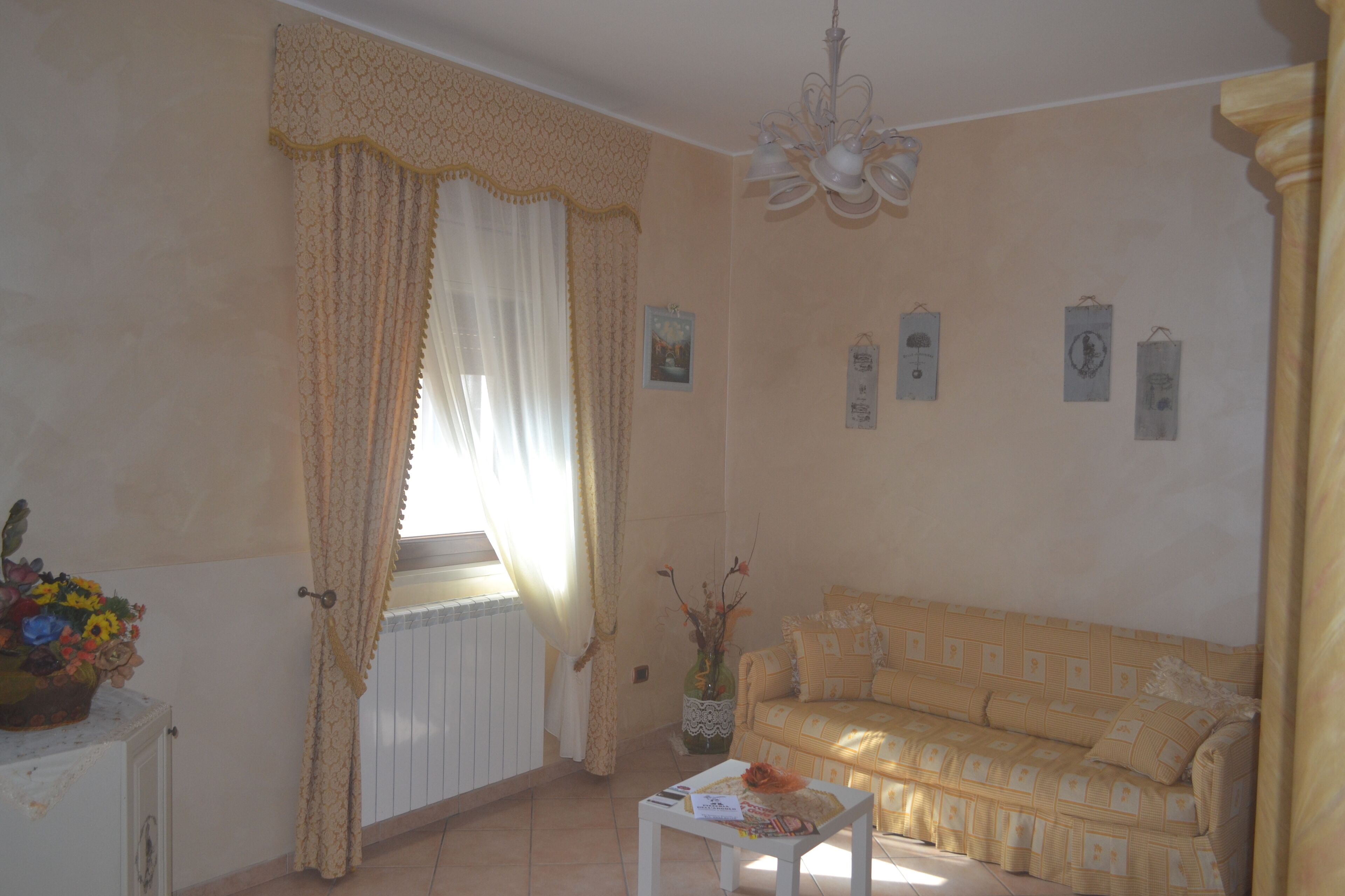 Ein Wohnzimmer mit einem beigen Sofa, einem weißen Tisch, einer Vase mit Blumen und einem Fenster mit Vorhängen.