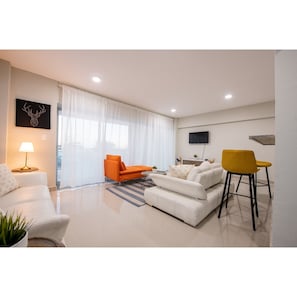 A modern living room with a white sofa, an orange chair, a yellow chair, a flat-screen TV, and a wall-mounted artwork. - Amazing Ocean view Unit Malecon (Santo Domingo)