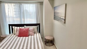 A bedroom with a bed, a window with sheer curtains, a bedside stool, and a framed picture on the wall. - Amazing Ocean view Unit Malecon (Santo Domingo)