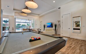 Ein Billardtisch mit Kugeln, ein Fernseher und ein Stativ in einem Raum mit großen Fenstern.