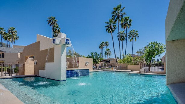 A swimming pool with a water feature, surrounded by palm trees and modern buildings. - Cozy Condo! New low rates for Spring/Summer 2026! Pet Friendly! (Tucson)