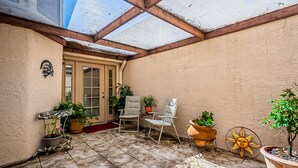 A sunlit patio with a wooden ceiling, two chairs, potted plants, and a door with shutters. - Cozy Condo! New low rates for Spring/Summer 2026! Pet Friendly! (Tucson)
