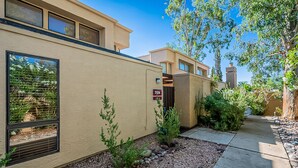 A modern building with a beige exterior, a numbered address sign, and a landscaped garden with small shrubs and a gravel pathway. - Cozy Condo! New low rates for Spring/Summer 2026! Pet Friendly! (Tucson)