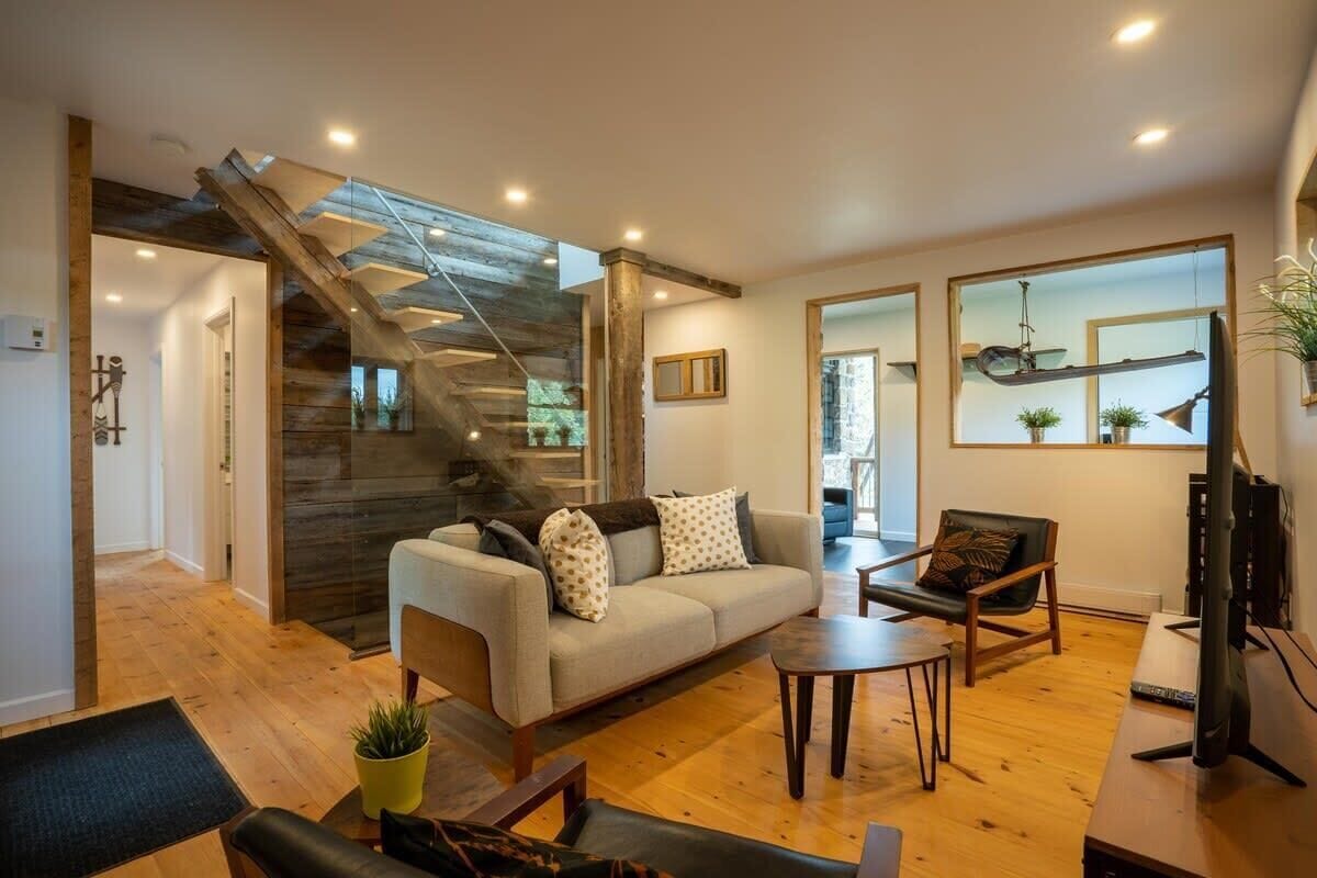 A modern living room with a sofa, coffee table, and chairs. A staircase with wooden accents leads to another level.