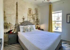 A bedroom with a large bed, a headboard with a cityscape mural, two bedside tables with lamps, and a window with curtains.