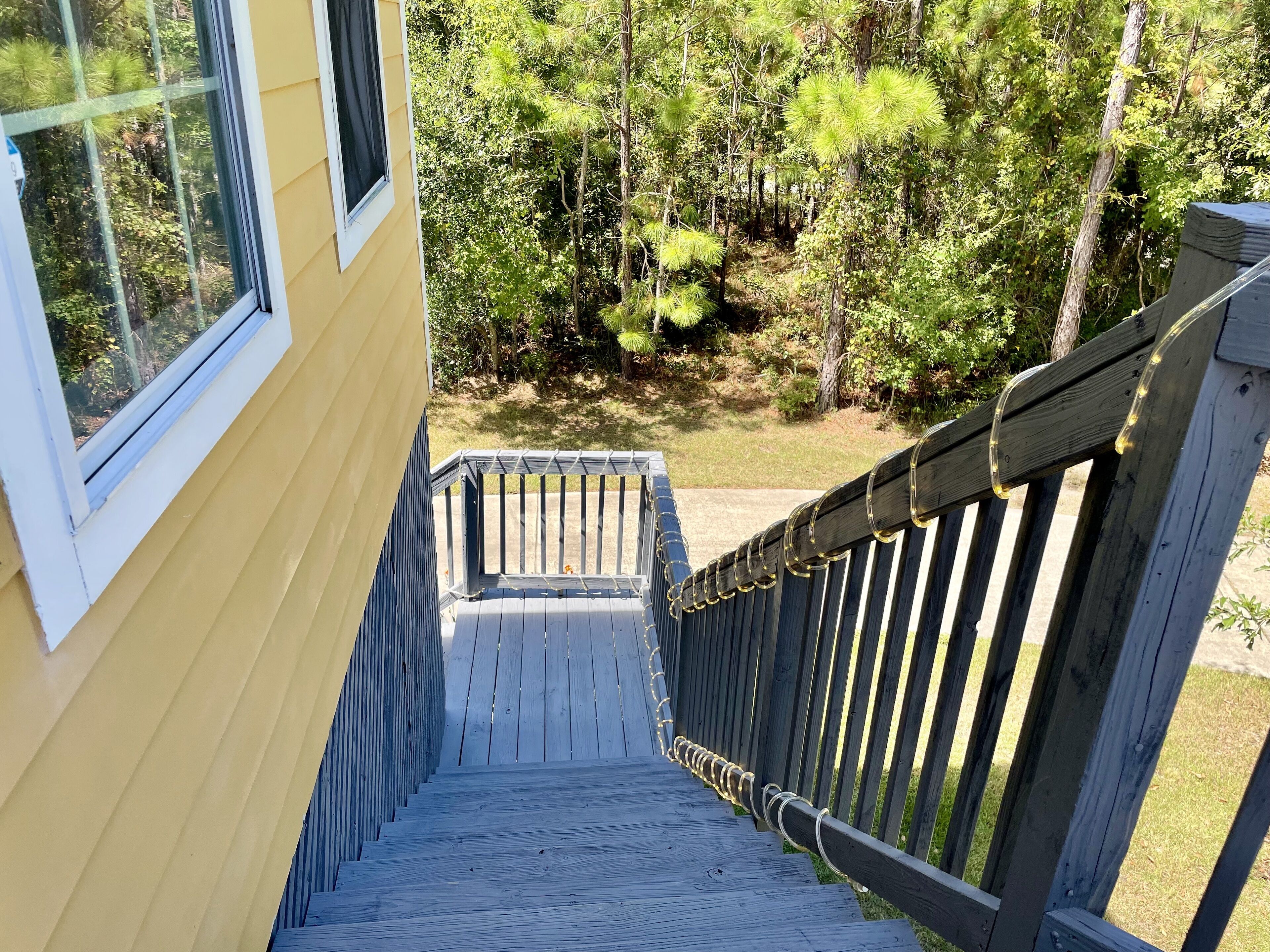 A wooden staircase leading to a deck with a railing, surrounded by trees and a yellow building.