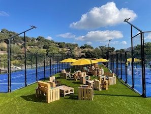 An outdoor seating area with wooden furniture and yellow umbrellas, surrounded by a pool and a fence.