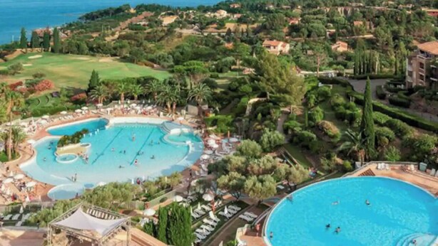 Un complejo turístico con una piscina grande central, una piscina más pequeña y una cancha de tenis.
