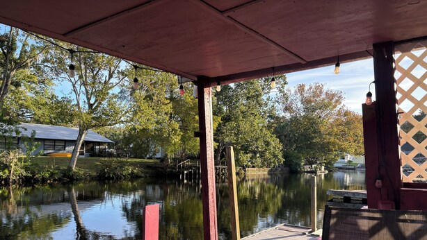 A wooden dock extending over a calm body of water, with a covered seating area and a view of trees and a building in the background. - Newly Remodeled Weeki Wachee River Retreat (Spring Hill)