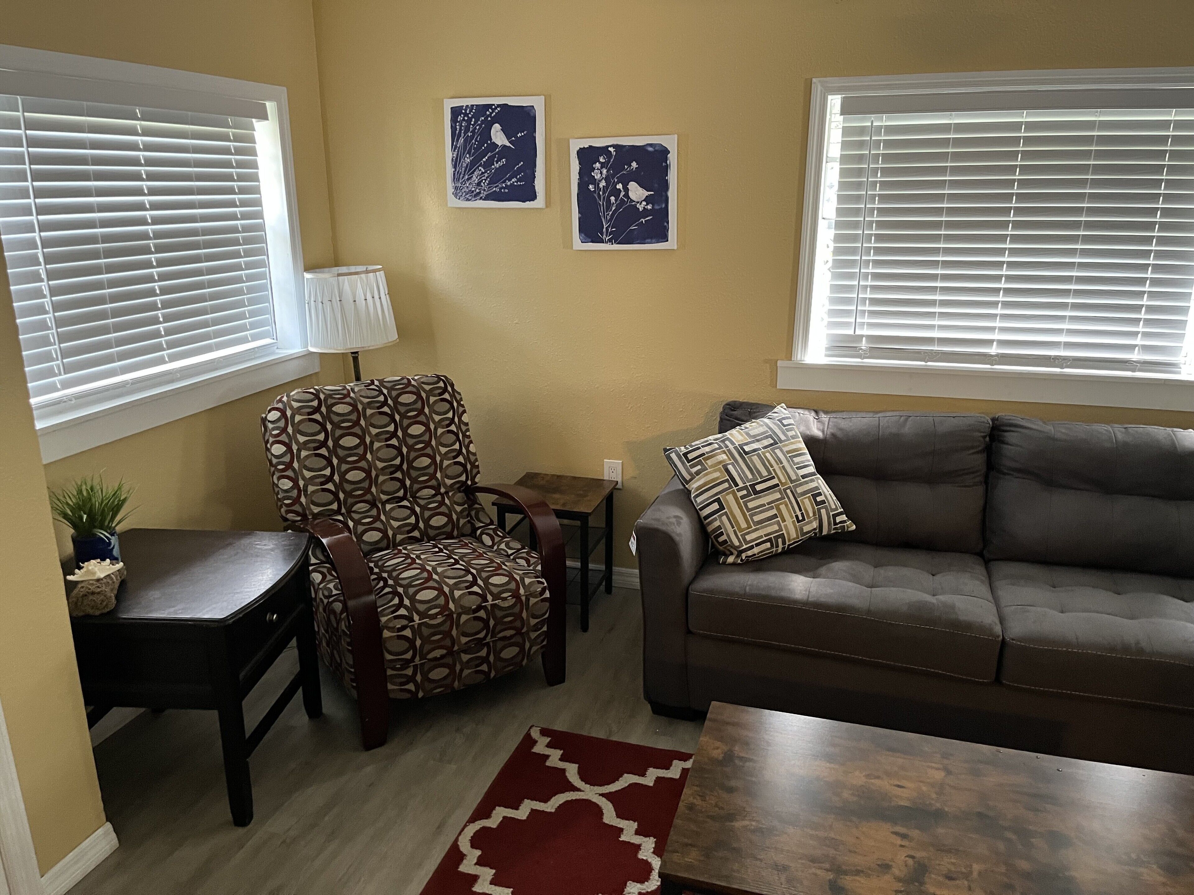 A living room with a patterned armchair, a brown sofa, a small table, and two wall art pieces.