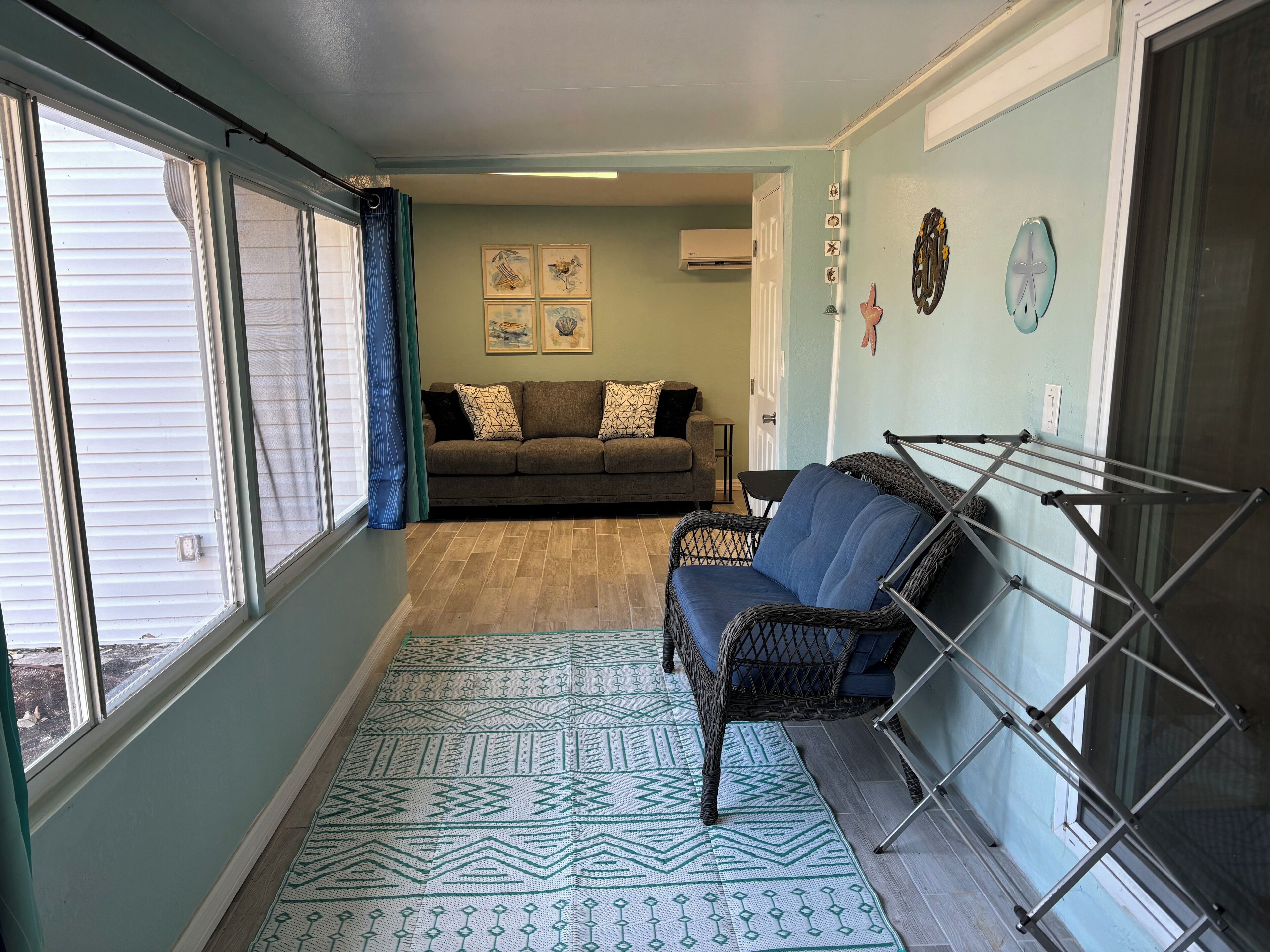 Une pièce comprenant un canapé, un fauteuil en osier, un tapis à motifs, un étendoir et un mur orné de tableaux encadrés.