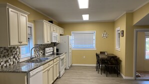 A compact kitchen with white cabinets, a granite countertop, and a built-in dishwasher. There is a dining area with a table and chairs, and a window with blinds. - Newly Remodeled Weeki Wachee River Retreat (Spring Hill)