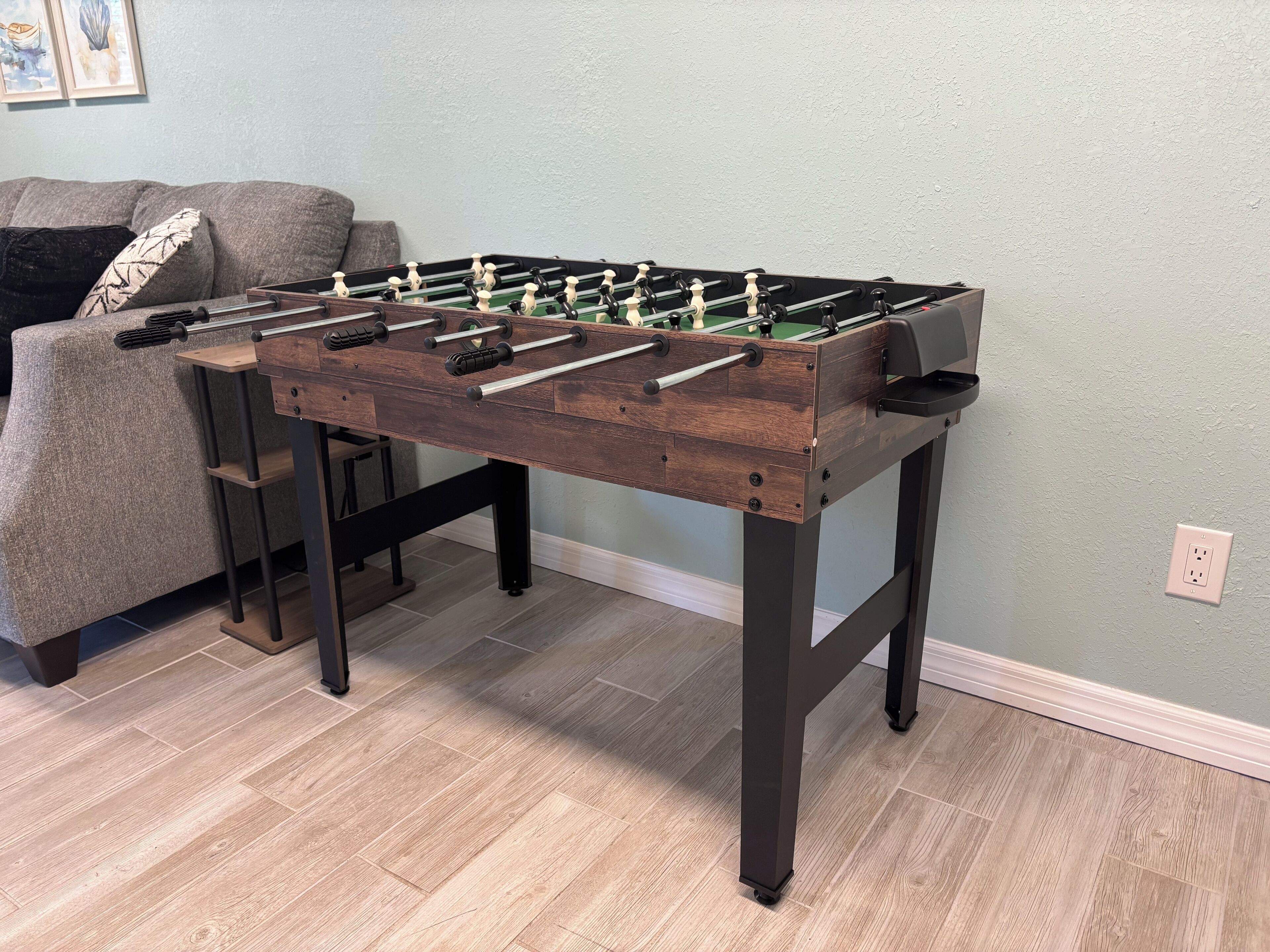 A wooden foosball table in a room with a gray couch and a light blue wall.