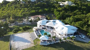 A large, white, multi-story house with a swimming pool, surrounded by lush greenery and a tennis court. - Luxury 5BD Villa - Massive Pool & Outdoor Vibe (Gros Islet)