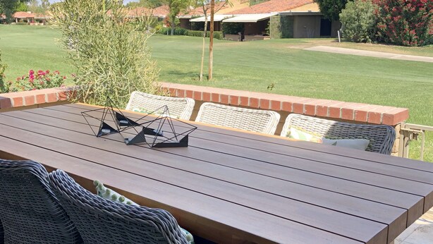 A wooden patio table with wicker chairs, a brick border, and a view of a well-maintained lawn and houses in the background. - Bright, Stylish Desert Condo with Mountain Views + Private Courtyard (Rancho Mirage)