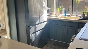 A modern kitchen with a large stainless steel refrigerator, dark blue cabinets, and a window with blinds. - Bright, Stylish Desert Condo with Mountain Views + Private Courtyard (Rancho Mirage)