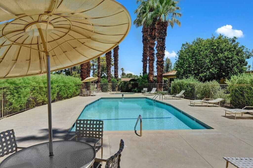 A rectangular swimming pool with a clear blue water surface, surrounded by lounge chairs and umbrellas, with palm trees and greenery in the background.