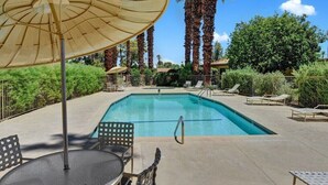 A rectangular swimming pool with a clear blue water surface, surrounded by lounge chairs and umbrellas, with palm trees and greenery in the background. - Bright, Stylish Desert Condo with Mountain Views + Private Courtyard (Rancho Mirage)
