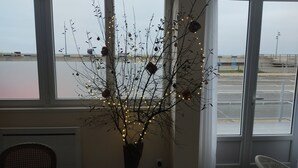 Une pièce avec une grande fenêtre, une plante en pot avec des branches et des guirlandes lumineuses, une chaise et une table.