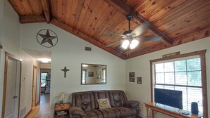 A living room with a brown leather couch, wooden ceiling, and a ceiling fan. There is a window with blinds and a sign that says "Hiking Trails". - Longhorn Lake House - Arcridgeranchresort.com (Ben Wheeler)