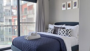 Une chambre d’hôtel moderne avec une grande fenêtre, un lit bien fait recouvert d’une couette bleu foncé, et une vue sur la ville.
