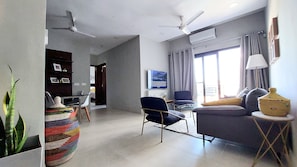 Uma sala de estar moderna com sofá, poltronas, mesa de centro e televisão. Há ventiladores de teto, uma janela com cortinas e um aparelho de ar condicionado.