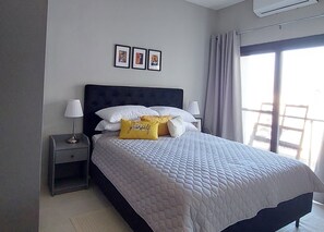 Cama bem arrumada com colcha acolchoada, duas luminárias de cabeceira e um criado-mudo com abajur. Três quadros emoldurados na parede acima da cama.