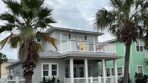 A two-story house with a balcony, surrounded by palm trees and a white picket fence. - 3 BR/3 BA Village Walk House, Close to the Beach, Community Pool! (Port Aransas)