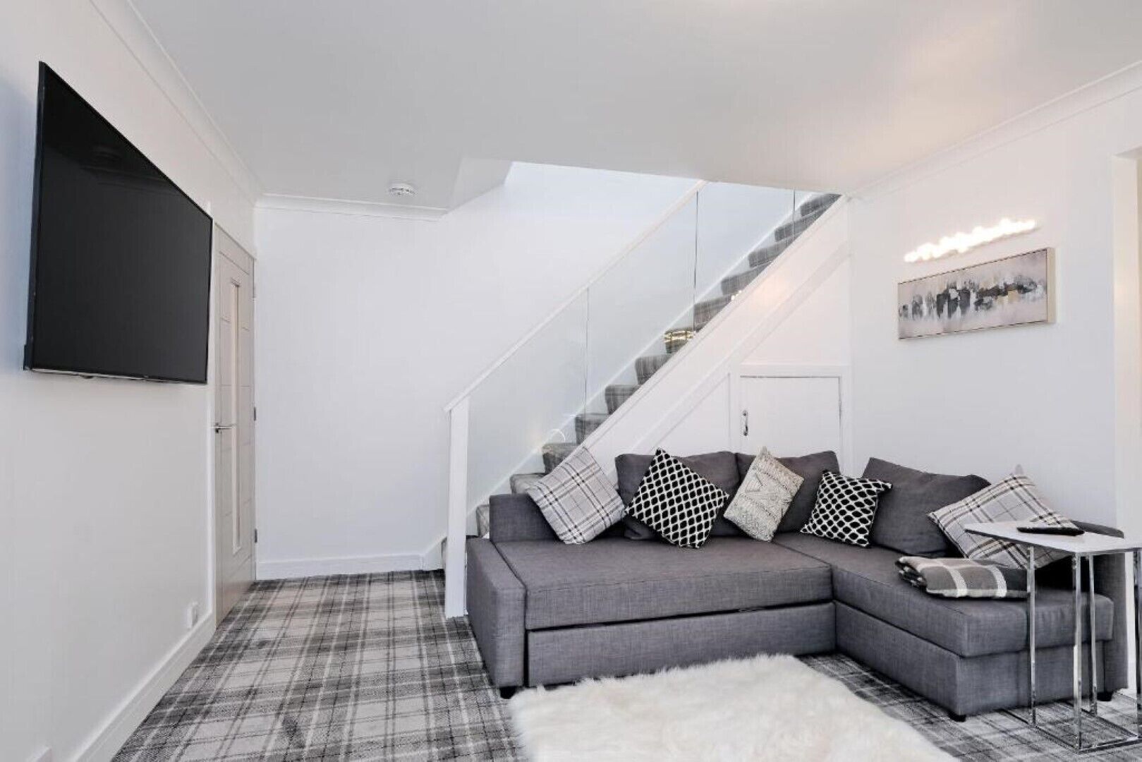 A modern living room with a grey sectional sofa, a plaid rug, a white fluffy rug, a small white side table, and a staircase with a glass railing.