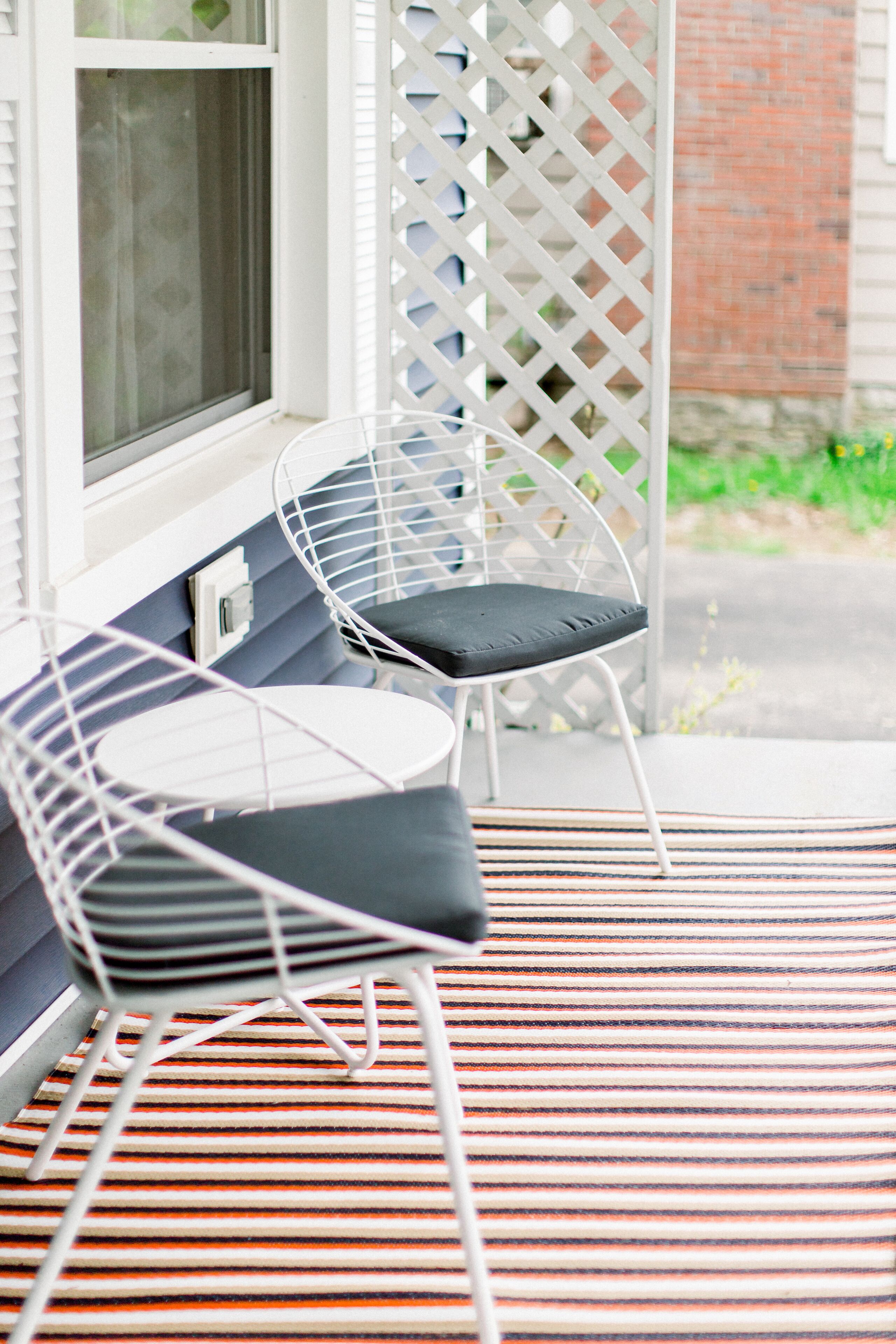 A porch with two white wire-framed chairs with dark cushions, a small round table, and a striped rug.