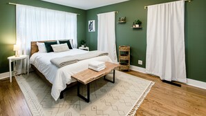 Un dormitorio con cama, mesitas de noche, un banco de madera, una alfombra y un estante en la pared con plantas.