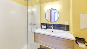 A bathroom with a glass shower enclosure, a white sink with a wooden vanity, a round mirror, and a yellow wall. - The Yellow Nest, Studio at Victoire Square (Burdeos)