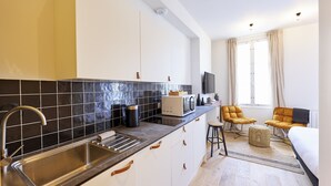 A modern kitchen with a black tiled backsplash, stainless steel sink, and white cabinets. There is a microwave, toaster, and a small table with chairs. The room has a large window with curtains and a bed with a black headboard. - The Yellow Nest, Studio at Victoire Square (Burdeos)