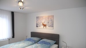 Quarto com cama, mesa de cabeceira, luminária na parede e pintura de um veado numa floresta nevada.