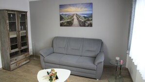 Uma sala de estar com um sofá cinzento, uma mesa de centro de vidro, um armário de madeira e uma pintura de uma paisagem com pôr do sol.