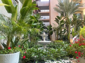 Stroll through a serene courtyard garden surrounded by lush tropical plants and vibrant flowers, with a central fountain and picturesque views from the multi-level balcony.