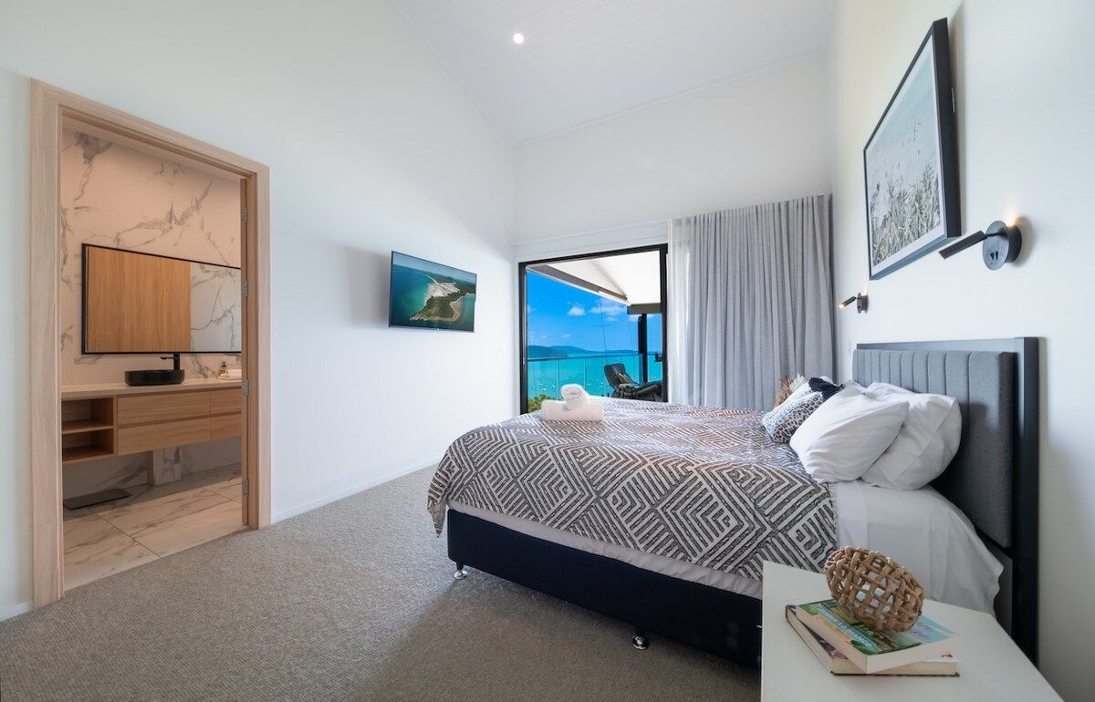A bedroom with a bed, a TV, a desk, and a view of the sea.