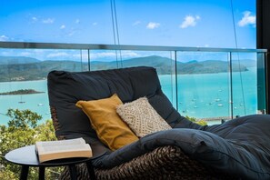 A balcony with a view of a turquoise sea and boats, a black cushioned chair, a yellow pillow, and a book on a small table. - Seven Islands (Airlie Beach)