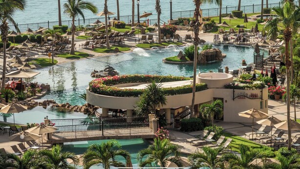Een resort met een groot zwembad, palmbomen en uitzicht op de oceaan.