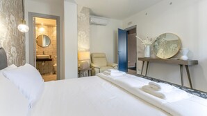 Habitación de hotel con cama, una silla, escritorio con espejo y baño visible a través de una puerta abierta.